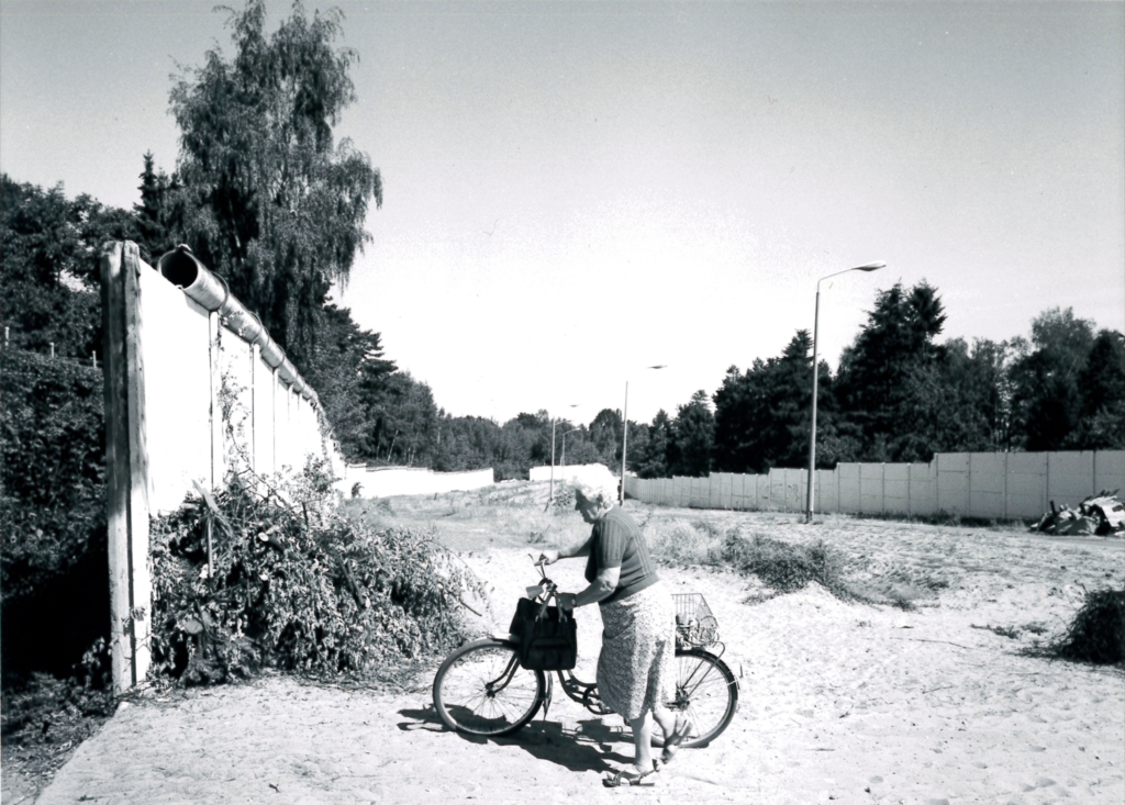 Schwarzweißfoto einer Frau, die ihr Fahrrad über den ehemaligen Grenzstreifen zwischen DDR und BRD schiebt.