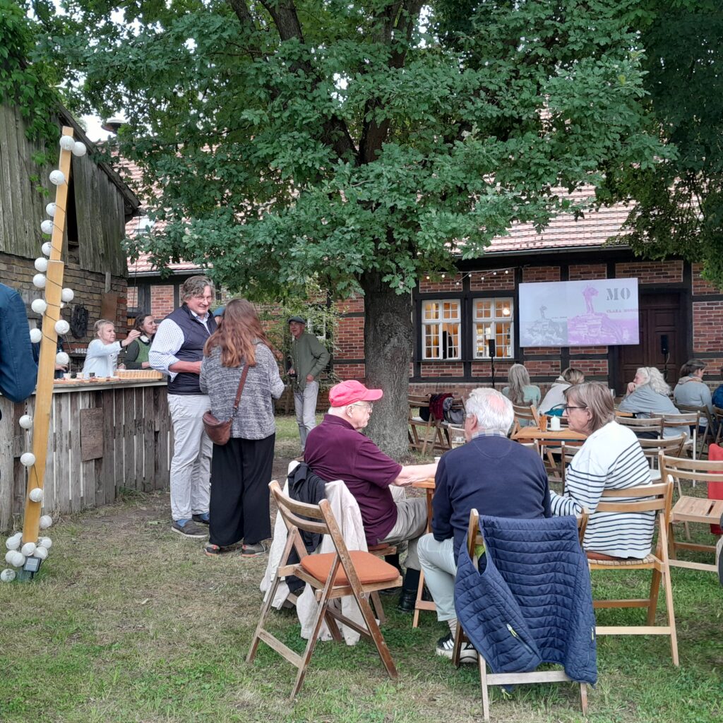 Mehrere Menschen unterhalten sich stehend und sitzen. Frau reicht Getränke hinter der Theke hervor.