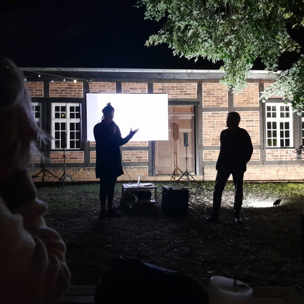 Zwei Menschen im Abenddunkel vor erleuchteter Leinwand. Frau spricht ins Mikrofon und redet gestenreich.
