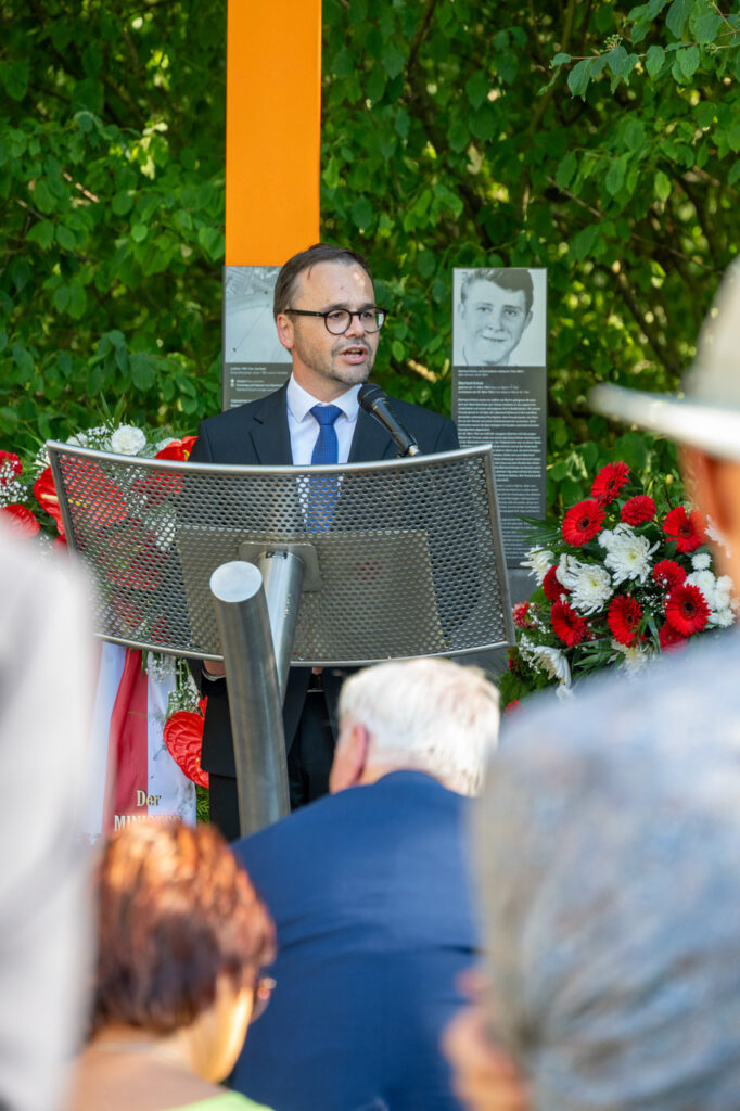 Mann hält an Pult Rede. Steht dabei vor Gedenkstele. Im Hintergrund außerdem Kränze zum Gedenken.