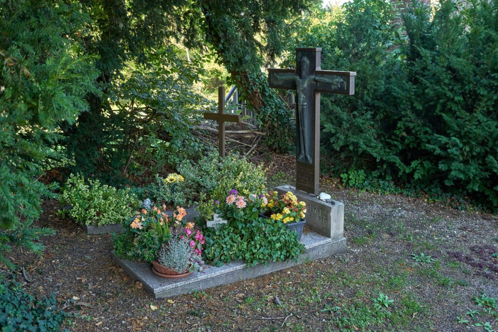 Grabstätte auf Friedhof. Großes Kreuz und viele Blumen. Waldlicher Charakter.
